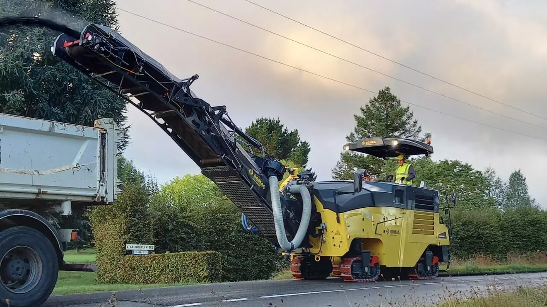 Bomag BM 1200 Cold Planer in operation on suburban road, unloading material into a dump truck