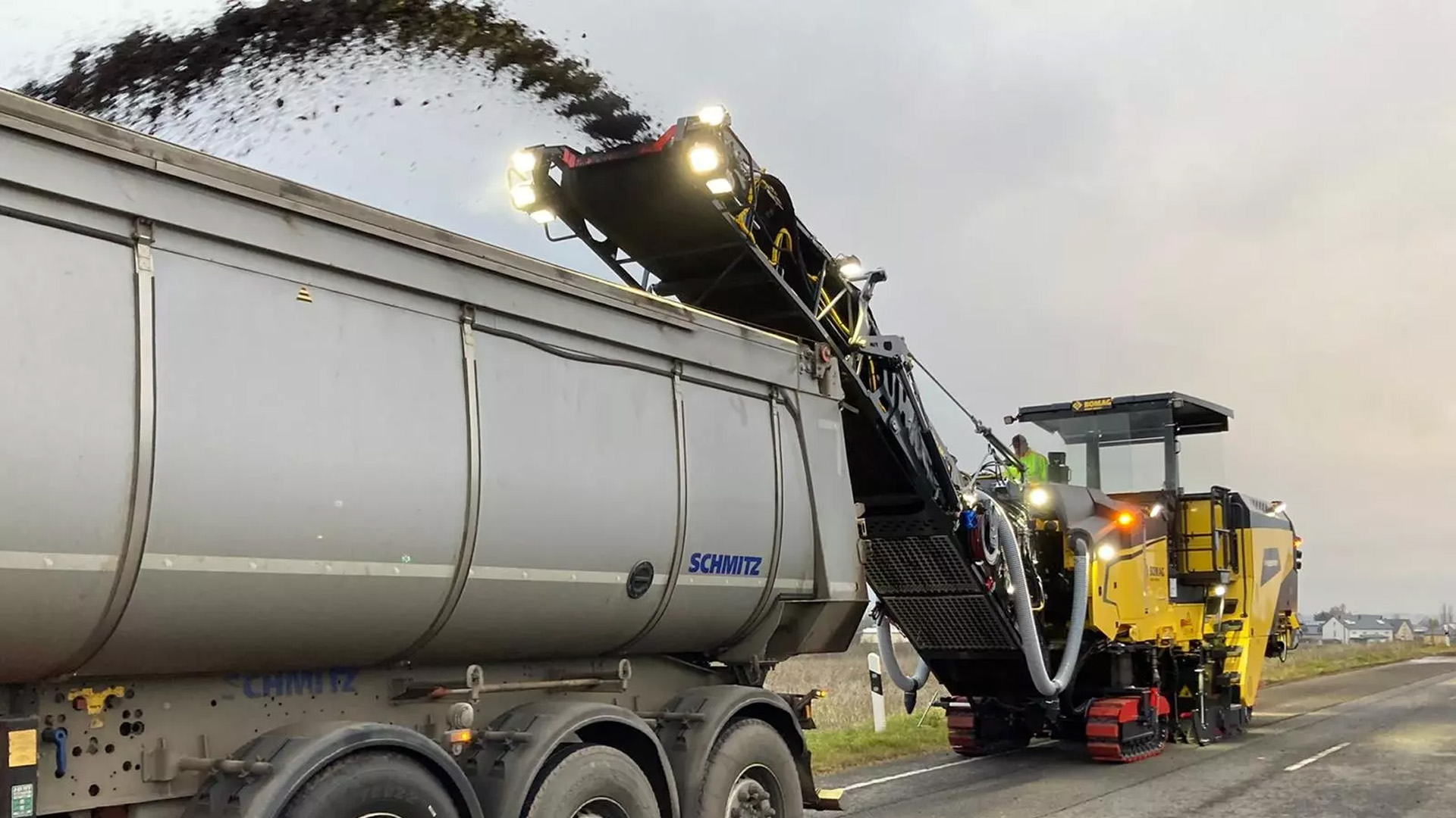 Bomag BM 2200 Milling Machine operating on a road, unloading material onto a dump truck with a worker in the operator's seat