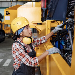 A woman wearing a hardhat and gloves works on the engine of a large yellow construction vehicle inside of a warehouse.