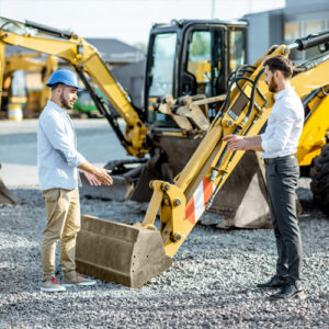 Builder with a sales consultant at the shop with heavy machinery. Two men with neatly styled hair stand facing each other near a large yellow excavator.
