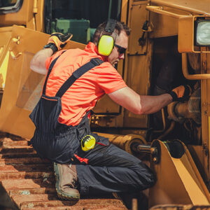 A mechanic wearing safety glasses and ear protection kneeling near a construction vehicle, inspecting the engine.