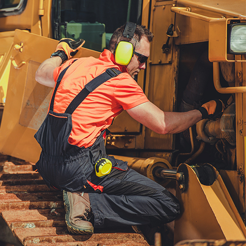 A mechanic wearing safety glasses and ear protection kneeling near a construction vehicle, inspecting the engine.